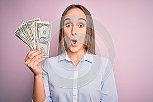 Young beautiful woman holding bunch of dollars banknotes over isolated pink background scared in shock with a surprise face,