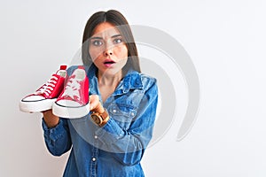 Young beautiful woman holding casual sneakers standing over isolated white background scared in shock with a surprise face, afraid