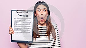 Young beautiful woman holding clipboard with contract document over pink background scared and amazed with open mouth for