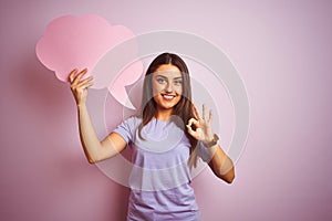 Young beautiful woman holding cloud speech bubble over isolated pink background doing ok sign with fingers, excellent symbol