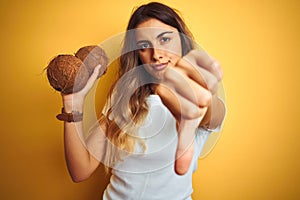 Young beautiful woman holding coconut over yellow isolated background with angry face, negative sign showing dislike with thumbs