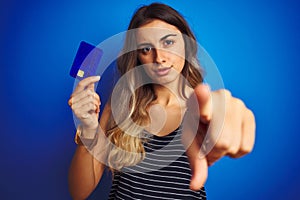 Young beautiful woman holding credit card over blue isolated background pointing with finger to the camera and to you, hand sign,