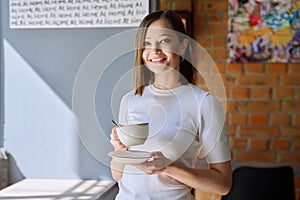 Young beautiful woman holding cup of coffee, looking out window, gray wall copy space