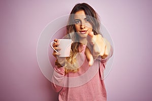 Young beautiful woman holding cup of coffee over pink isolated background pointing with finger to the camera and to you, hand