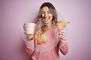 Young beautiful woman holding cup of coffee over pink isolated background pointing and showing with thumb up to the side with