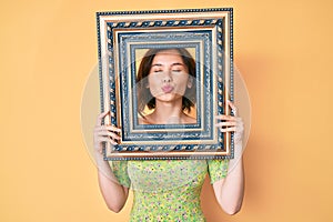 Young beautiful woman holding empty frame looking at the camera blowing a kiss being lovely and sexy