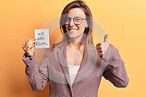Young beautiful woman holding we are equal paper smiling happy and positive, thumb up doing excellent and approval sign