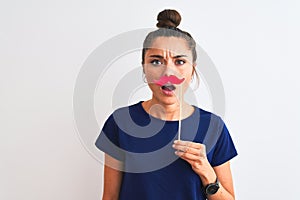 Young beautiful woman holding fanny party mustache over isolated white background scared in shock with a surprise face, afraid and