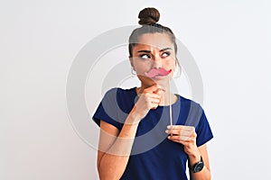 Young beautiful woman holding fanny party mustache over isolated white background serious face thinking about question, very