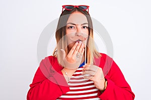 Young beautiful woman holding fanny party mustache standing over isolated white background cover mouth with hand shocked with