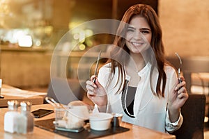 Young beautiful woman holding fork and knife looks hungry ready to eat meat burger with fries for lunch in trendy cafe eating
