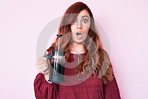 Young beautiful woman holding french coffee maker scared and amazed with open mouth for surprise, disbelief face