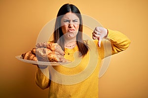 Young beautiful woman holding french croissant pastry over yellow background with angry face, negative sign showing dislike with