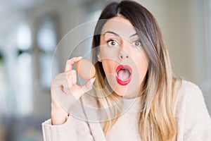 Young beautiful woman holding fresh egg at home scared in shock with a surprise face, afraid and excited with fear expression
