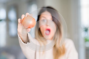 Young beautiful woman holding fresh onion at home scared in shock with a surprise face, afraid and excited with fear expression