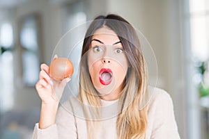 Young beautiful woman holding fresh onion at home scared in shock with a surprise face, afraid and excited with fear expression