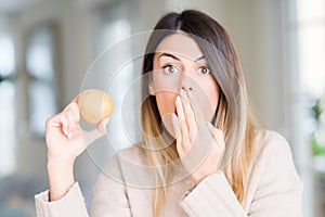 Young beautiful woman holding fresh potato at home cover mouth with hand shocked with shame for mistake, expression of fear,