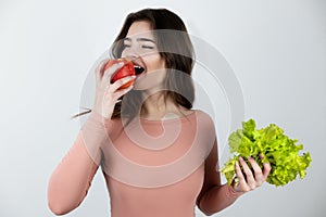 Young beautiful woman holding green salad in one hand and biting fresh apple in another looking happy being on diet isolated white
