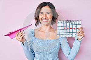 Young beautiful woman holding heart calendar and paper airplane smiling with a happy and cool smile on face