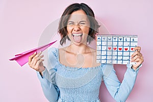 Young beautiful woman holding heart calendar and paper airplane sticking tongue out happy with funny expression