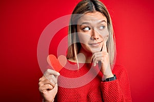Young beautiful woman holding heart card standing over isolated red background serious face thinking about question, very confused