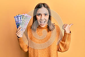 Young beautiful woman holding indian rupee banknotes pointing thumb up to the side smiling happy with open mouth