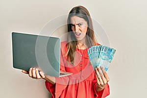 Young beautiful woman holding laptop and holding brazilian real in shock face, looking skeptical and sarcastic, surprised with