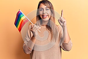 Young beautiful woman holding lgtbq flag smiling with an idea or question pointing finger with happy face, number one