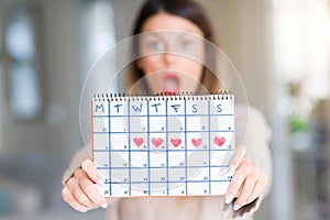 Young beautiful woman holding menstruation calendar at home scared in shock with a surprise face, afraid and excited with fear