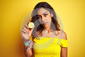 Young beautiful woman holding middle lemon standing over isolated yellow background with a confident expression on smart face