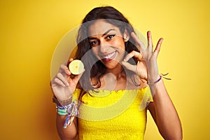 Young beautiful woman holding middle lemon standing over isolated yellow background doing ok sign with fingers, excellent symbol