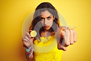 Young beautiful woman holding middle lemon standing over isolated yellow background pointing with finger to the camera and to you,