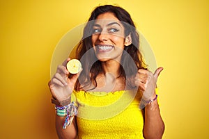 Young beautiful woman holding middle lemon standing over isolated yellow background pointing and showing with thumb up to the side