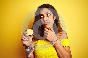 Young beautiful woman holding middle lemon standing over isolated yellow background serious face thinking about question, very