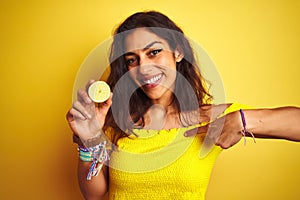 Young beautiful woman holding middle lemon standing over isolated yellow background with surprise face pointing finger to himself
