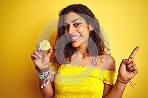 Young beautiful woman holding middle lemon standing over isolated yellow background very happy pointing with hand and finger to