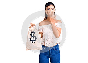 Young beautiful woman holding money bag with dollar symbol covering mouth with hand, shocked and afraid for mistake