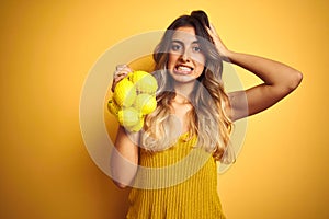 Young beautiful woman holding net of lemons over yellow  background stressed with hand on head, shocked with shame and