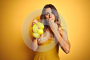 Young beautiful woman holding net of lemons over yellow isolated background cover mouth with hand shocked with shame for mistake,