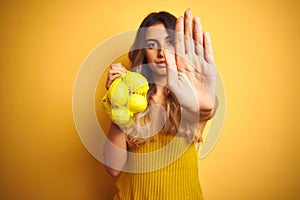 Young beautiful woman holding net of lemons over yellow isolated background with open hand doing stop sign with serious and