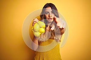 Young beautiful woman holding net of lemons over yellow isolated background pointing with finger to the camera and to you, hand