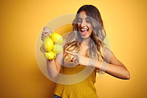 Young beautiful woman holding net of lemons over yellow isolated background very happy pointing with hand and finger