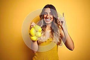 Young beautiful woman holding net of lemons over yellow isolated background very happy pointing with hand and finger to the side