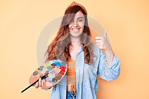 Young beautiful woman holding paintbrush and palette smiling happy and positive, thumb up doing excellent and approval sign