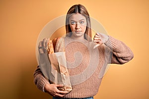 Young beautiful woman holding paper bag with bread over isolated yellow background with angry face, negative sign showing dislike