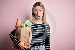 Young beautiful woman holding paper bag with purchase over isolated pink background scared in shock with a surprise face, afraid