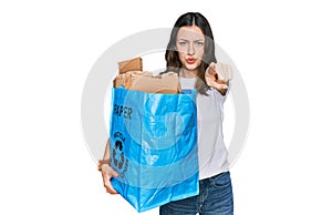 Young beautiful woman holding recycling wastebasket with paper and cardboard pointing with finger to the camera and to you,