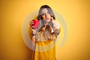 Young beautiful woman holding  red cup of coffee over yellow isolated background pointing with finger to the camera and to you,