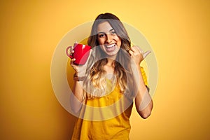 Young beautiful woman holding  red cup of coffee over yellow isolated background pointing and showing with thumb up to the side