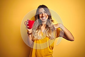 Young beautiful woman holding  red cup of coffee over yellow isolated background with surprise face pointing finger to himself